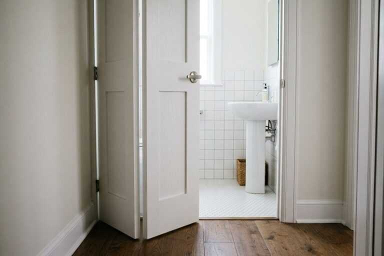 Open white double door revealing a bright, modern bathroom with tiled walls and a pedestal sink.